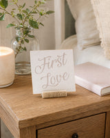 First Love 4x4 Christian Shelf Sitter in a natural wood stand on a warm nightstand with a candle, greenery, and pale textiles.
